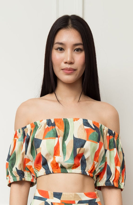 Woman wearing a colorful off-shoulder top and skirt set against a white paneled wall.